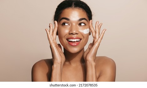 Happy smiling woman applies facial moisturizing cream, nourish her skin, stands over brown background