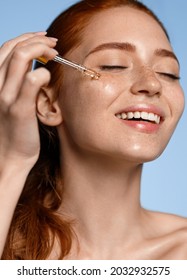 Happy smiling woman applying serum on her face, close eyes with pleasure. Redhead girl holding dropper with skin care product for healthy and glowing skin tone, blue background - Shutterstock ID 2032932575