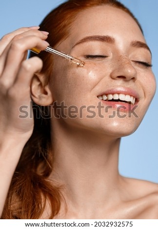 Happy smiling woman applying serum on her face, close eyes with pleasure. Redhead girl holding dropper with skin care product for healthy and glowing skin tone, blue background
