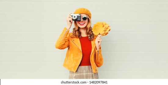 happy smiling woman with autumn yellow maple leaves, retro camera taking picture in french beret over gray wall background