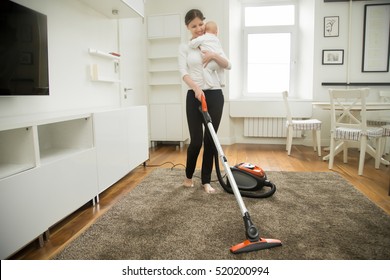 Happy smiling woman doing vacuum cleaning the carpet in the living room holding a child, modern interior. Busy mum. Home, housekeeping concept.