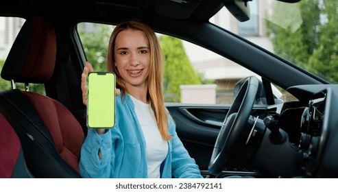 Happy smiling woman driver sitting behind the wheel of premium modern car showing smartphone with vertical green screen. Green screen, blank phone, application. Chroma key smartphone technology - Powered by Shutterstock