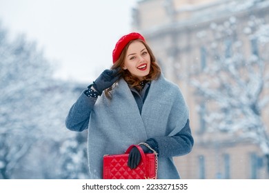 Happy smiling woman enjoying winter holidays, walking in snow covered street of European city. Model wearing red beret, grey scarf, coat, gloves, holding quilted bag. Copy, empty space for text  - Powered by Shutterstock