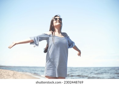 Happy smiling woman in free bliss on ocean beach standing with open hands. Portrait of a brunette female model in summer dress enjoying nature during travel holidays vacation outdoors