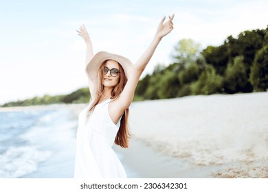 Happy smiling woman in free happiness bliss on ocean beach standing with a hat, sunglasses, and rasing hands. Portrait of a multicultural female model in white summer dress enjoying nature during trav