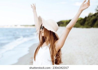 Happy smiling woman in free happiness bliss on ocean beach standing with a hat, sunglasses, and rasing hands. Portrait of a multicultural female model in white summer dress enjoying nature during trav