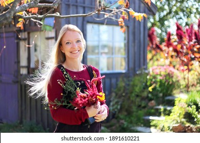 A Happy, Smiling Woman Gardener Is Holding Flowers Outside Her Cottage Garden And Shed.