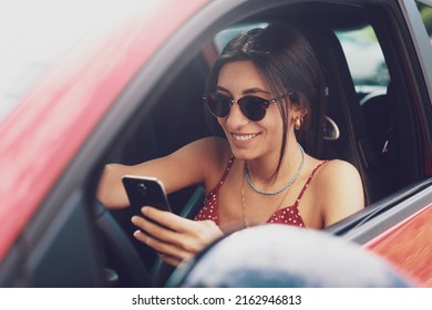 Happy Smiling Woman Holding Her Smart Phone Sitting Inside A Car. Female Driver Laughing Reading Message And Chatting In Mobile.