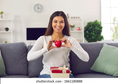 Happy Smiling Woman In Love Sitting On Sofa At Home With Valentine Present Box On Lap, Holding Red Heart And Saying Thank You For Gifts. Portrait Of Girlfriend For Skype, Zoom Or FaceTime Video Call