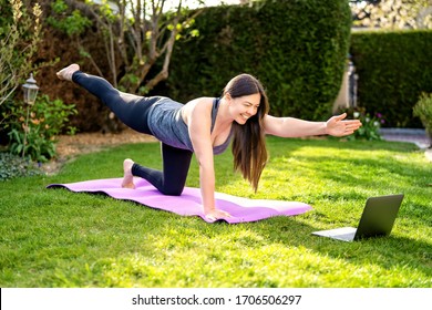 Happy smiling woman practicing pilates lesson online in garden outdoors during quarantine. Doing sport at home following guide or online tutorial or trainer instructions on laptop. 