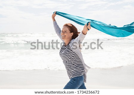 Happy smiling woman running with lightblue scarf enjoying at beach. Beautiful woman holding blue fabric at wind with copy space. Middle aged mature woman having fun, freedom and carefree concept.