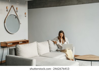 Happy smiling woman sitting on couch and using laptop at living room at home, watching funny video, learning language, video calling, mother working online drink coffee - Powered by Shutterstock