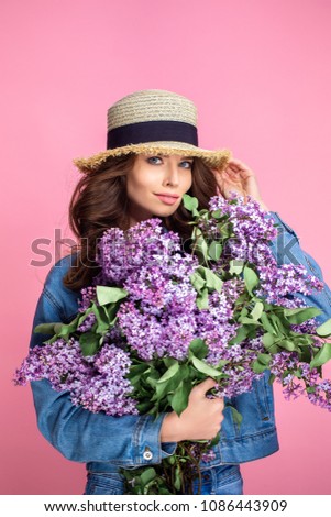 Happy smiling woman in straw hat posing with bouquet of lilac flowers over colorful pink background