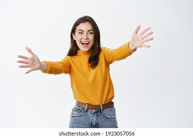 Happy smiling woman stretch out hands to greet guest, wants to hug or welcome you. Beautiful female model reaching arms to hold or receive something, standing against white background. - Powered by Shutterstock