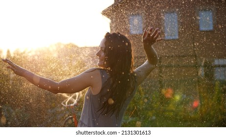 Happy Smiling Woman Is Under The Rain. Stunning Golden Sun Rays Shine On Playful Young Woman Enjoying A Summer Rain. Slow Motion