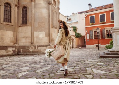 Happy smiling woman wearing straw hat, long trench coat, holding basket with flowers, walking in street of European city. Lifestyle, travel concept. Full-length portrait. Copy, empty space for text