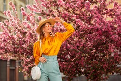 Happy Smiling Woman Wearing Trendy Straw Hat, Yellow Satin Blouse, Blue Trousers, Cat Eye Sunglasses, With Wicker Bag, Posing In Street Near Pink Spring Blossom Trees. Copy, Empty Space For Text 