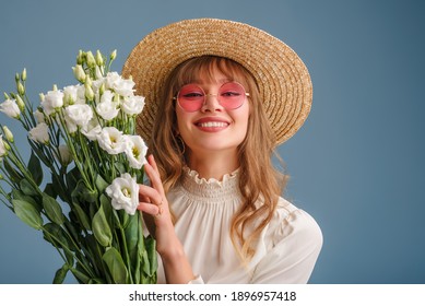 Happy Smiling Woman Wearing Trendy Spring Outfit: Pink Sunglasses, Straw Hat, Holding Flowers, Posing On Blue Background. Copy, Empty Space For Text