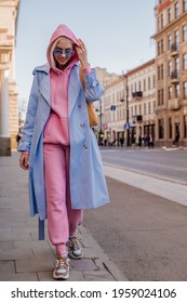 Happy Smiling Woman Wearing Trendy Sport Chic Style Outfit Walking In City. Streetstyle, Street Fashion Conception. Blue Trench Coat, Sunglasses, Pink Hoodie, Trousers, Sneakers, Yellow Bag