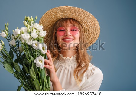Happy smiling woman wearing trendy spring outfit: pink sunglasses, straw hat, holding flowers, posing on blue background. Copy, empty space for text