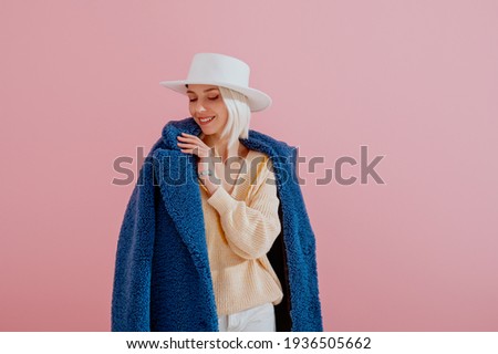 Happy smiling woman wearing trendy blue faux fur coat, yellow sweater, stylish silver wrist watch, white hat, posing on pink background. Fashion conception. Copy, empty space for text
