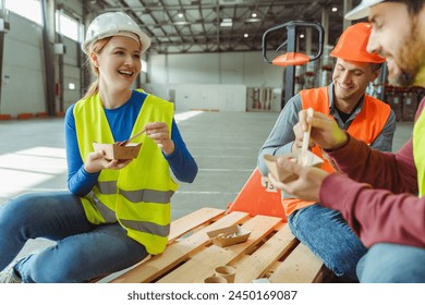 Happy, smiling workers, factory managers wearing hard hats, vests eating meal, sitting, talking while having lunch together in warehouse. Food concept - Powered by Shutterstock