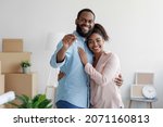 Happy smiling young african american loving family hugging in new home among boxes and showing house keys, man and woman spouses feel joy. Moving, relocating to own apartment, rental and real estate