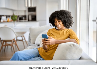 Happy smiling young African American woman customer sitting at home on sofa looking at cellphone holding mobile cell phone in hands texting using smartphone scrolling media, buying online. Authentic. - Powered by Shutterstock