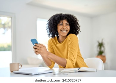 Happy smiling young African American woman customer sitting at home table and looking away while holding mobile cell phone using smartphone shopping online in app on cellphone in living room. - Powered by Shutterstock