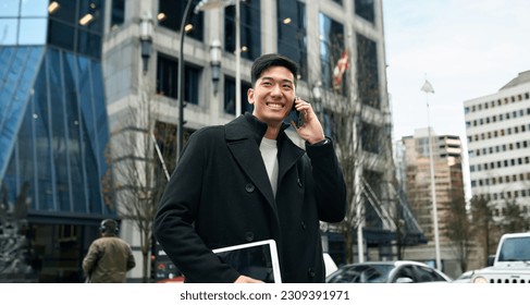 Happy smiling young Asian business man professional holding cellphone walking on big city urban street making business call, talking on cellular phone standing in office downtown.