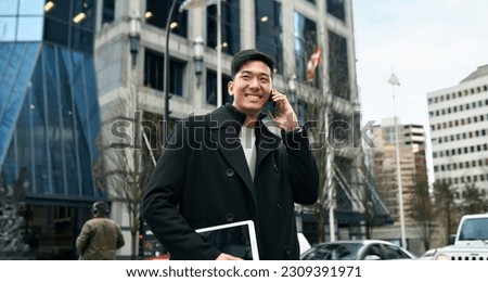 Happy smiling young Asian business man professional holding cellphone walking on big city urban street making business call, talking on cellular phone standing in office downtown.