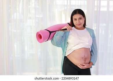 Happy Smiling Young Asian Pregnant Stnding On Floor While Holding Pink Mat For Yoga And Excercise Workout In Living Room At Home. Expectant Mother Prepare For Baby Birth During Pregnancy Concept