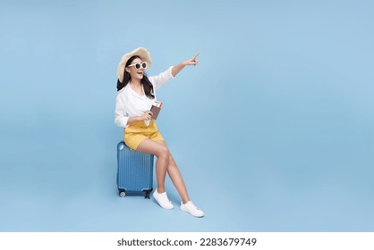 Happy smiling young Asian tourist woman sitting on luggage holding passport and hand pointing to copy space going to travel on holidays isolated on blue studio background. - Shutterstock ID 2283679749