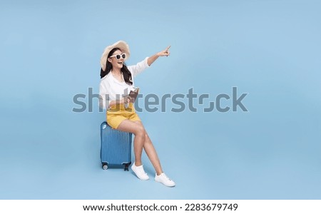 Happy smiling young Asian tourist woman sitting on luggage holding passport and hand pointing to copy space going to travel on holidays isolated on blue studio background.