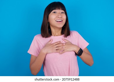 Happy Smiling Young Asian Woman Wearing Pink T-shirt Against Blue Background Has Hands On Chest Near Heart. Human Emotions, Real Feelings And Facial Expression Concept.