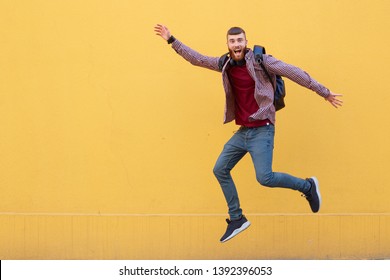 Happy smiling young attractive ginger bearded man jumping, wearing in basic clothes with backpack. Looking at the camera with wide open mouth over a yellow wall with copy space. - Shutterstock ID 1392396053