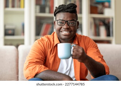 Happy smiling young black man in casual outwear wearing eyeglasses sitting on couch in living room, holding mug, african american guy enjoying morning coffee at home, copy space - Powered by Shutterstock