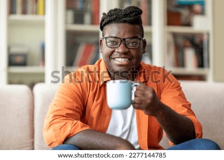 Happy smiling young black man in casual outwear wearing eyeglasses sitting on couch in living room, holding mug, african american guy enjoying morning coffee at home, copy space