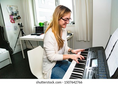 Happy smiling young blond Latin woman in glasses and casual clothes sitting on white chair at home, playing music on electronic piano. Education, holidays, weekend, leisure activities and hobby