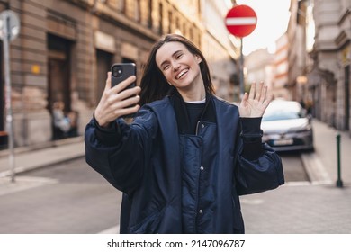 Happy smiling young blonde woman making video call outdoors in city. Stylish female traveler taking selfie or video for internet blog. Girl wear navy blue bomber jacket on the street.