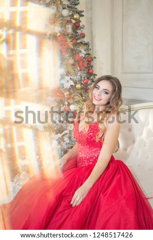 Happy smiling young blonde women in a long red evening dress sitting near Christmas tree with presents in the room in a room decorated for xmas. Beautiful New Year and Christmas scene.