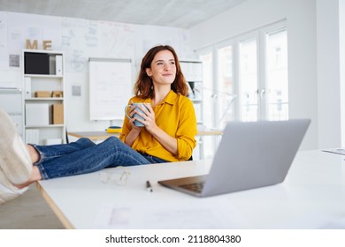 Happy smiling young businesswoman sitting daydreaming in the office as she relaxes with a mug of coffee and her feet up during a morning break - Powered by Shutterstock