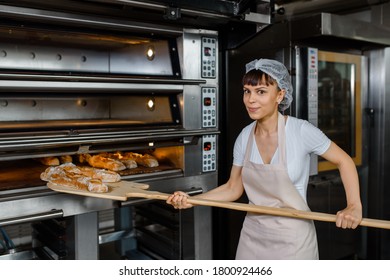 Happy smiling young caucasian woman baker is taking off from oven the french baguette bread with wood peel at baking manufacture factory and looking to the camera. French baguette bread bakery  - Shutterstock ID 1800924466