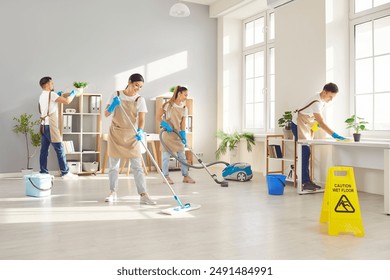 Happy smiling young cleaners men and women in uniform wiping desk, mopping floor and vacuuming. Cheerful team of professional janitors washing office workplace. Cleaning service concept.