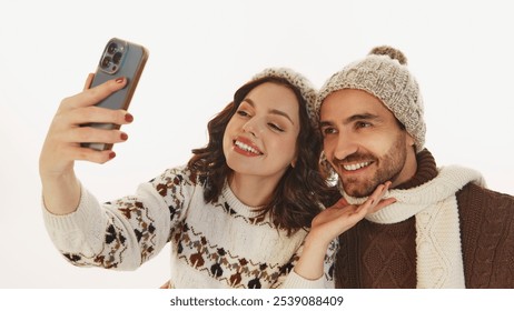 Happy, smiling young couple, man and woman dressed in cozy winter clothes taking selfie isolated on white background. Warm memories. Concept of winter, vacation, holiday, leisure activity
