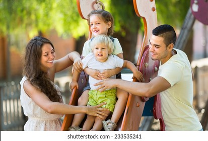 Happy Smiling Young Family Of Four At Children's Playground In Park