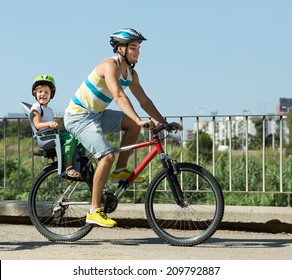 Happy Smiling Young Father On Bicycle With Child In Baby Seat Behind 