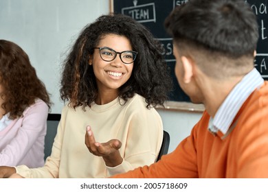 Happy Smiling Young Female African American Woman Student Talking To Male Indian Coworker. Creative Team Group Talking Brainstorming Discussing Startup Project Sitting At Desk In Classroom Office.