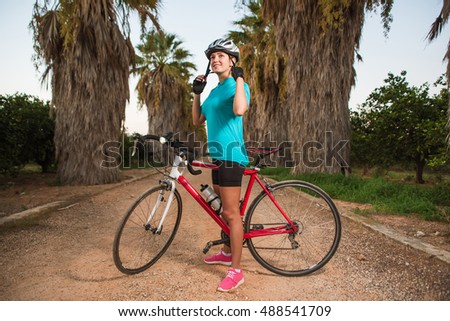 Happy smiling young female cyclist wearing helmet standing with bike on path with palms in sunset. Nature, healthy and active lifestyle concept