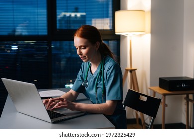 Happy Smiling Young Female Doctor In Blue Green Medical Uniform Working Typing On Laptop Computer Sitting At Desk In Dark Hospital Room Near Window At Night.